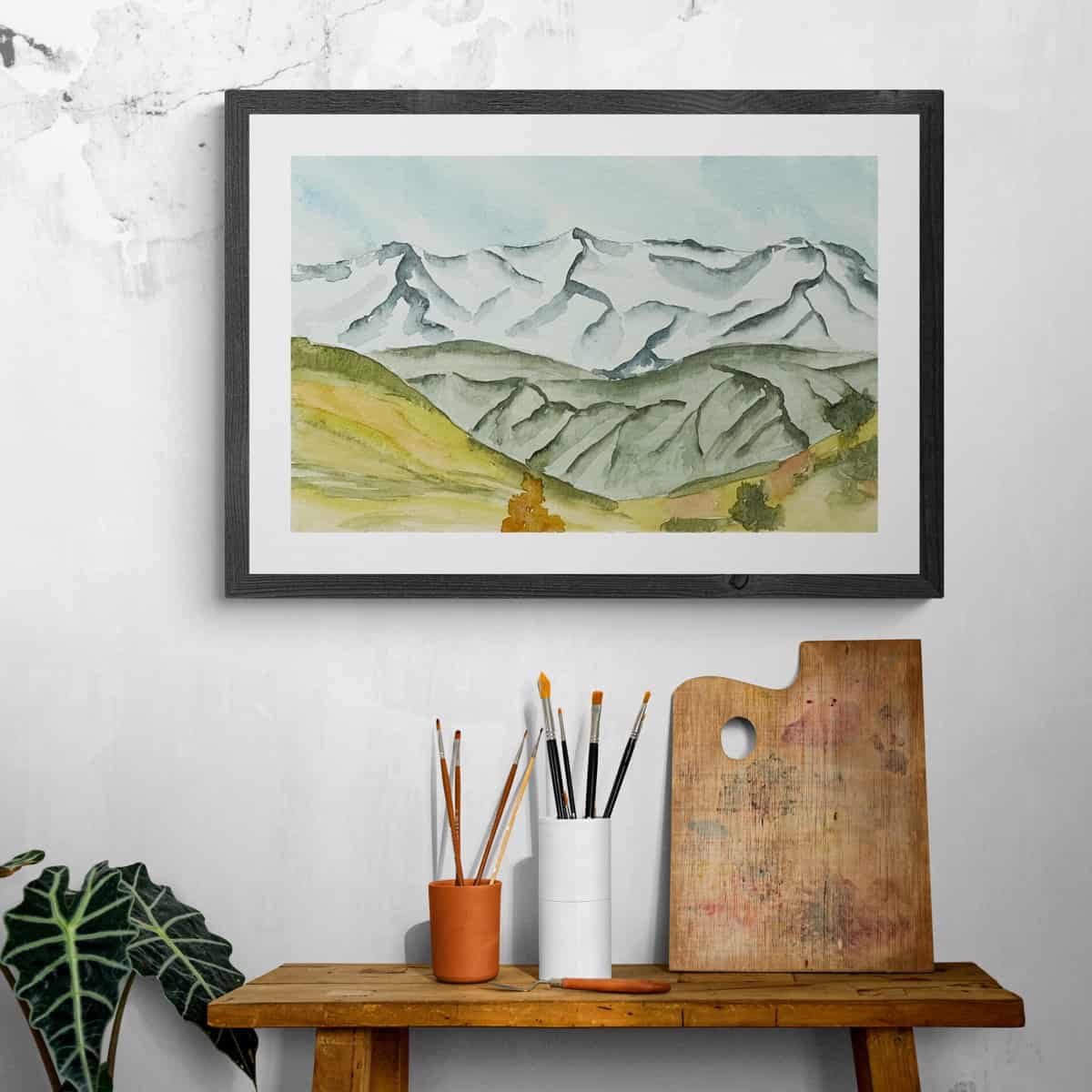 Framed painting of a mountain scene hanging on a wall above an artists work table.