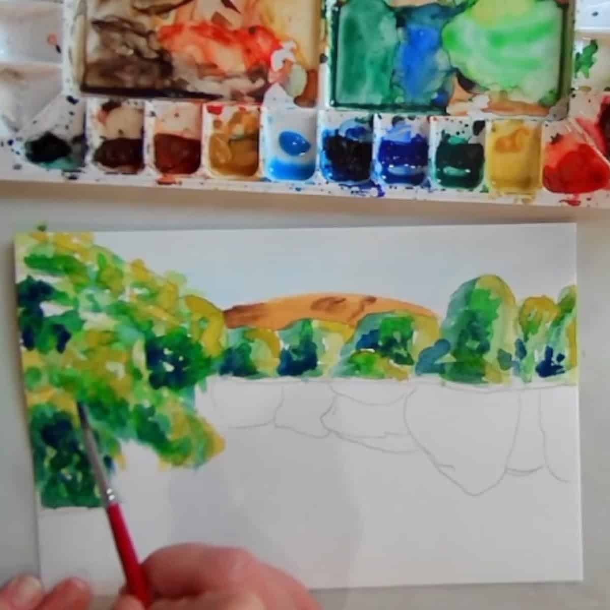 A lake scene with trees painted next to a watercolor palette. 