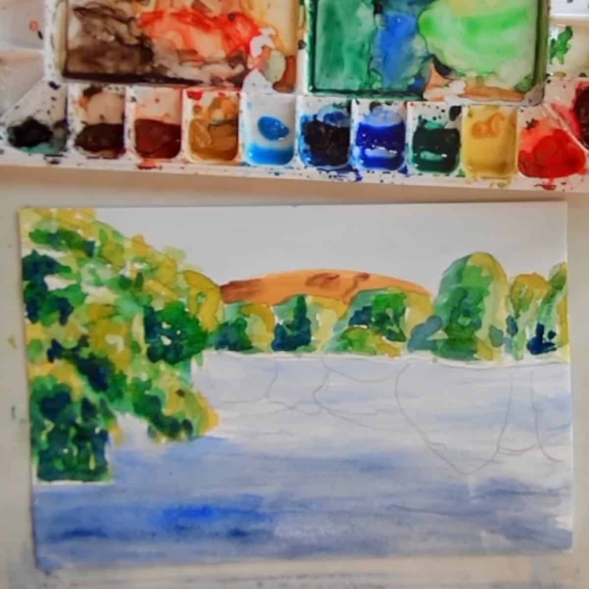 A lake scene with trees and water painted next to a watercolor palette. 