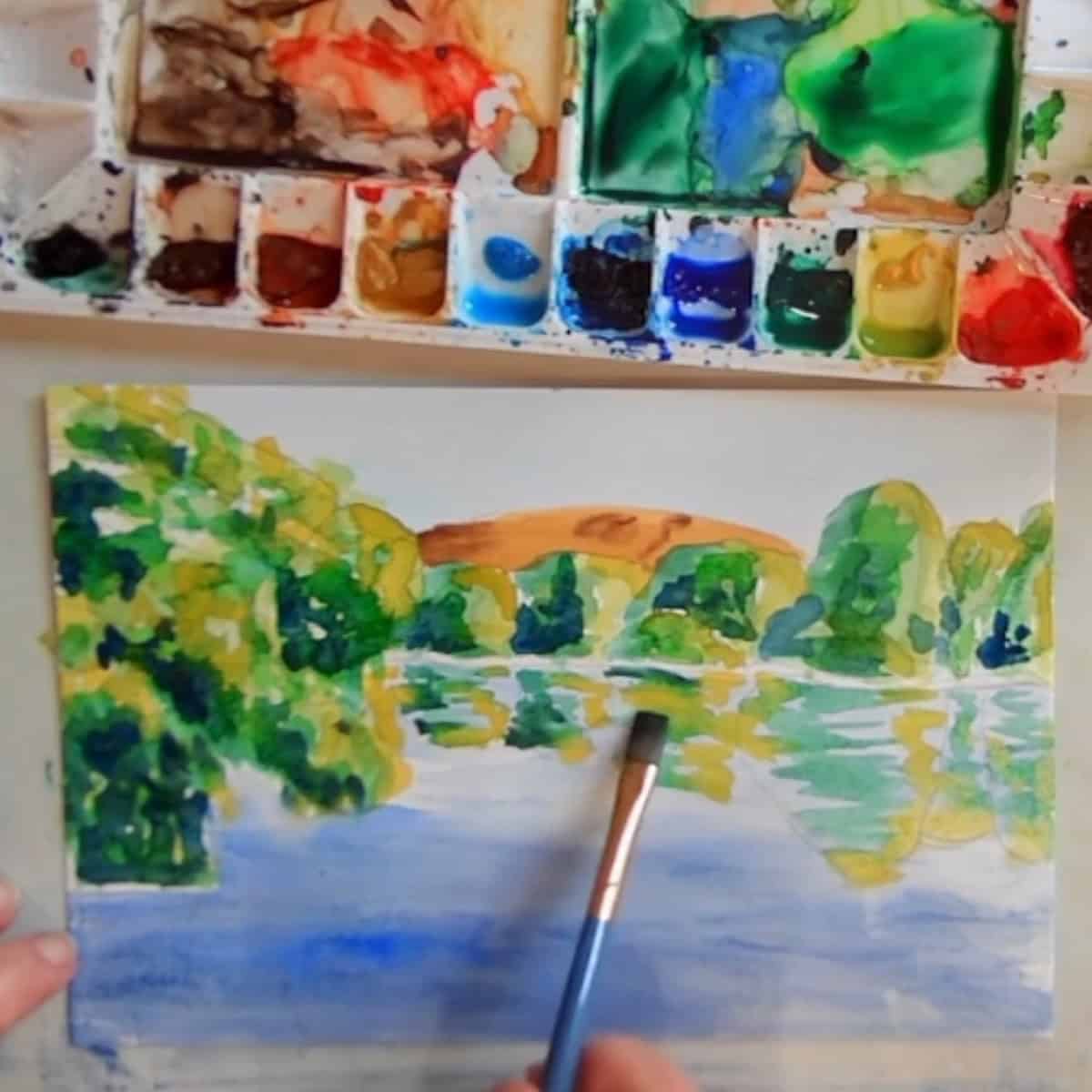 A lake scene with trees, water and reflections painted next to a watercolor palette. 