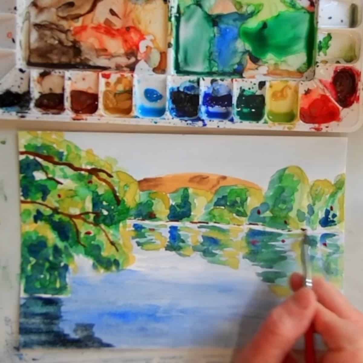 A lake scene with trees, water and reflections and artist's hand painting in details next to a watercolor palette. 