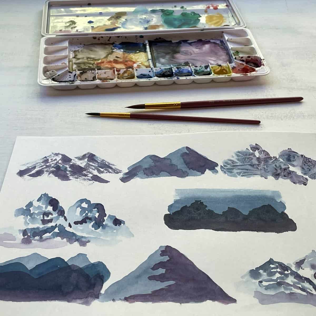 Up close painting of different watercolor mountains nect to paintbrushes and a paint pallette.