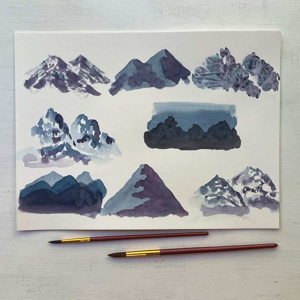 Watercolor painting of eight different styles of mountains next to some paintbrushes.