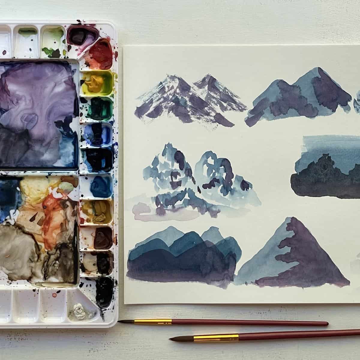 Up close painting of different watercolor mountains nect to paintbrushes and a paint pallette.