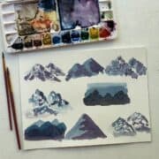 Watercolor paintings of 8 different styles of mountains next to a watercolor pallette and paintbrushes.