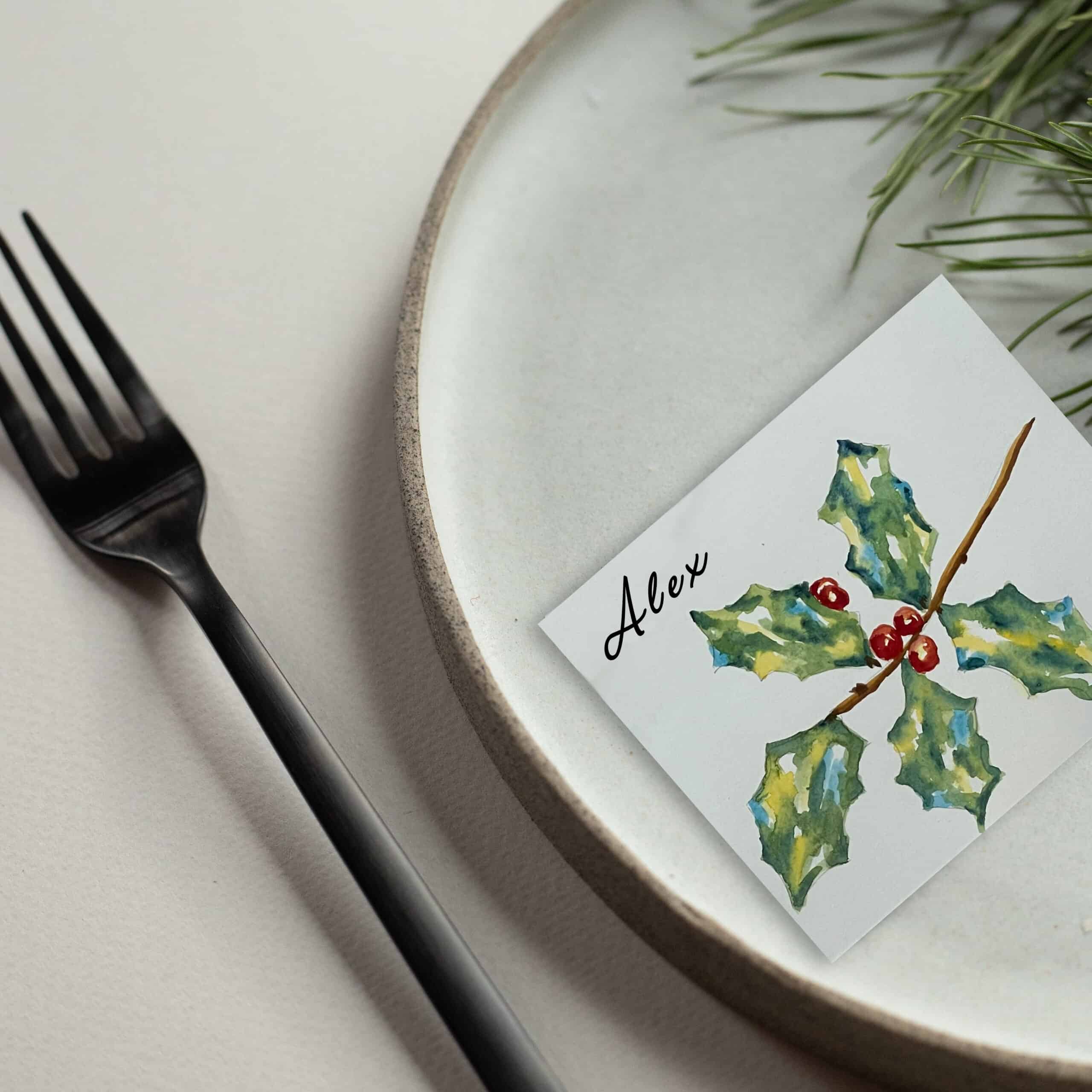 A name place setting painted with watercolor holly on a plate next to a fork. 