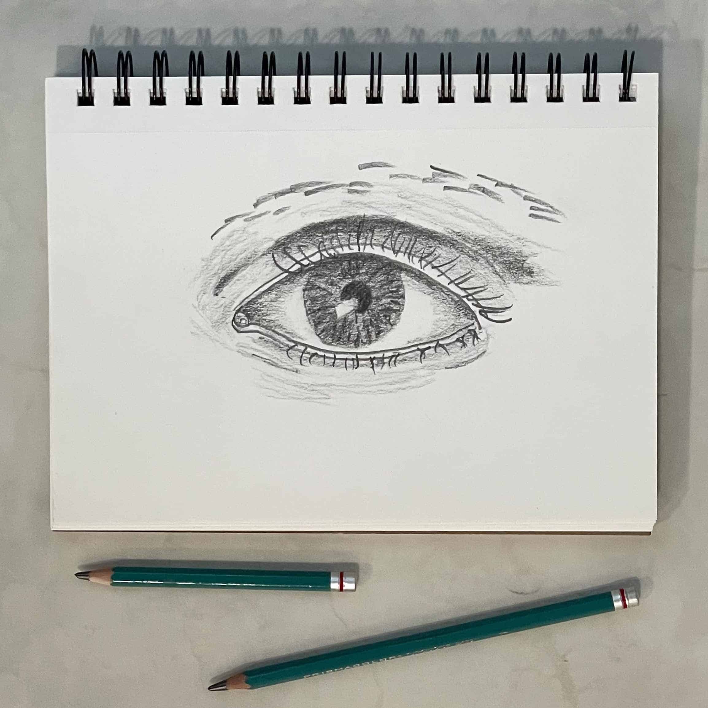 Pencil sketch of an eye on a sketch pad with drawing pencils.