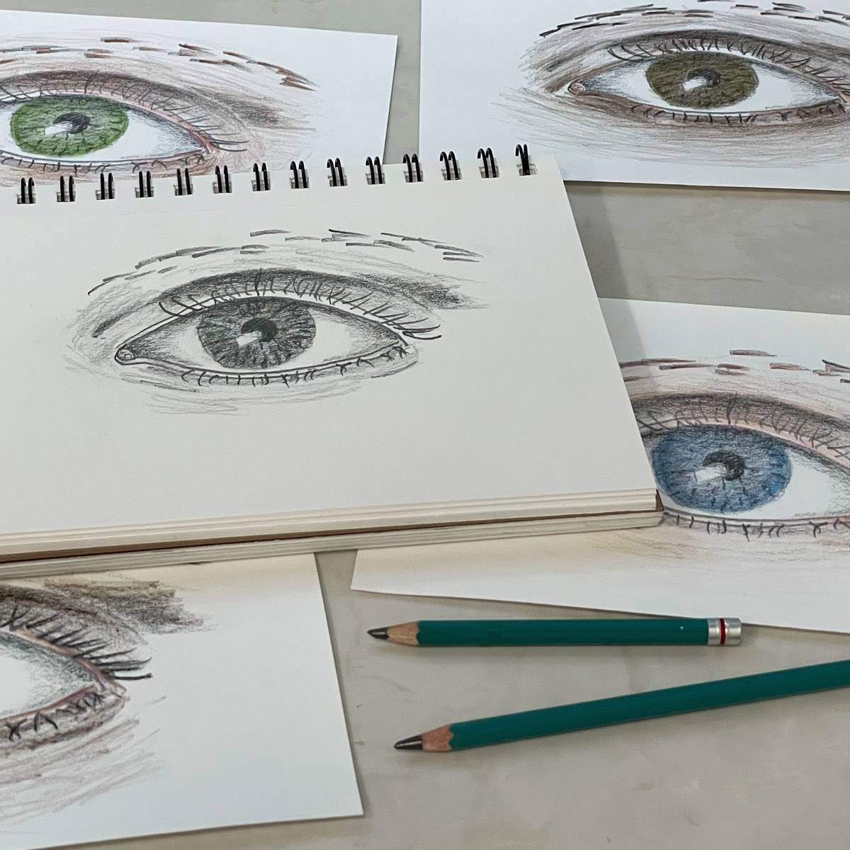 A pencil drawing of an eye on a sketchbook with drawing pencils next to colored pencil drawing of eyes.