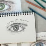 A pencil drawing of an eye on a sketchbook with drawing pencils next to colored pencil drawing of eyes.