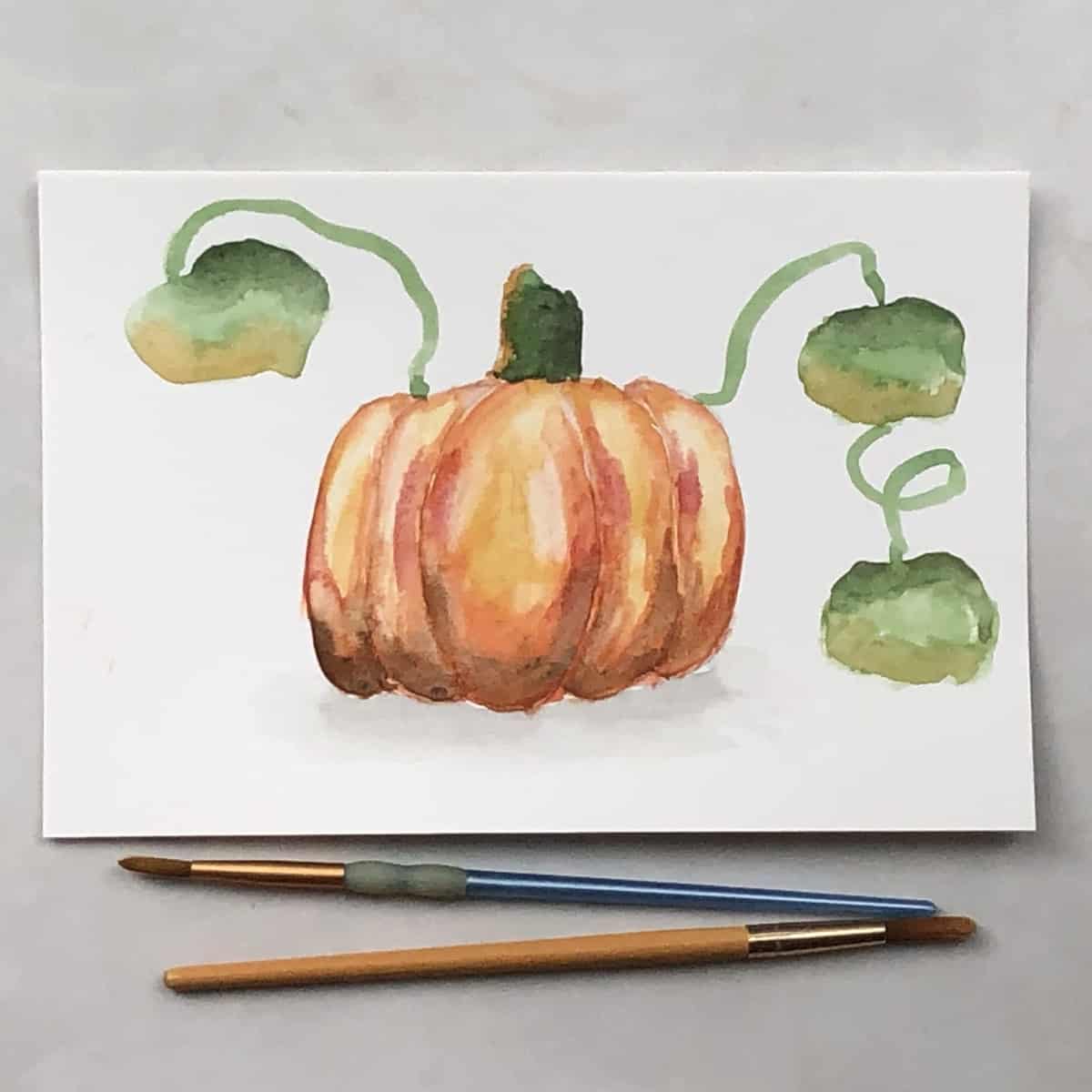 Painting of a pumpkin in watercolor paints next to paint brushes.