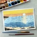 Watercolor painting of a beach sunset next to paint brushes and watercolor paints.