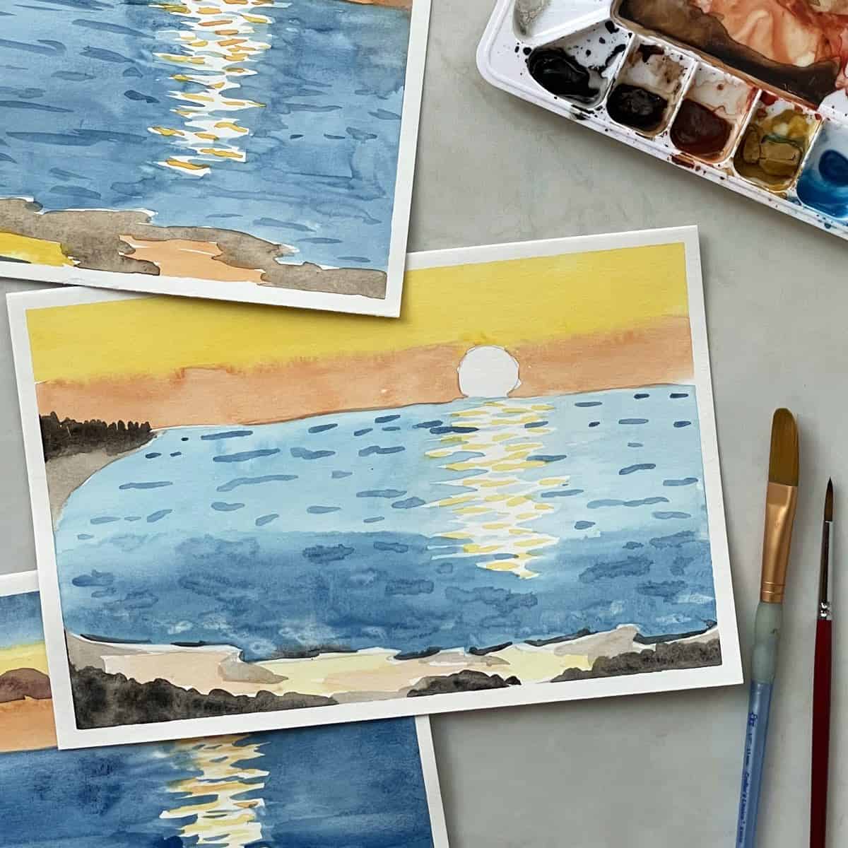 Watercolor paintings of a beach sunset next to paint brushes and watercolor paints.