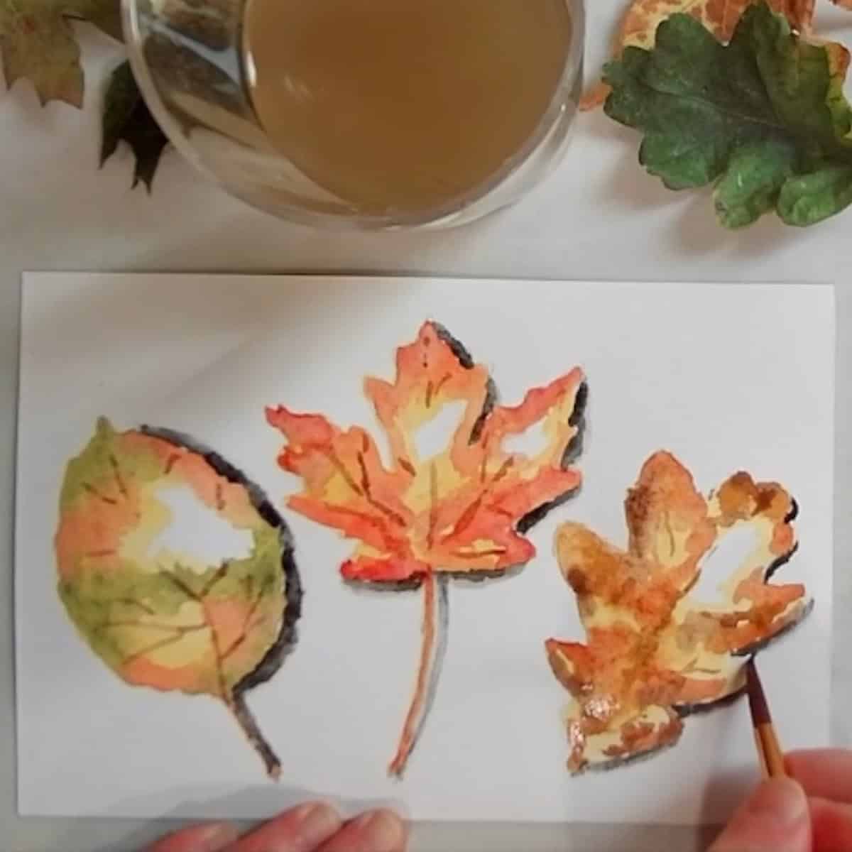 An artist painting shadows on the side of a colorful autumn leaf in watercolor paint.