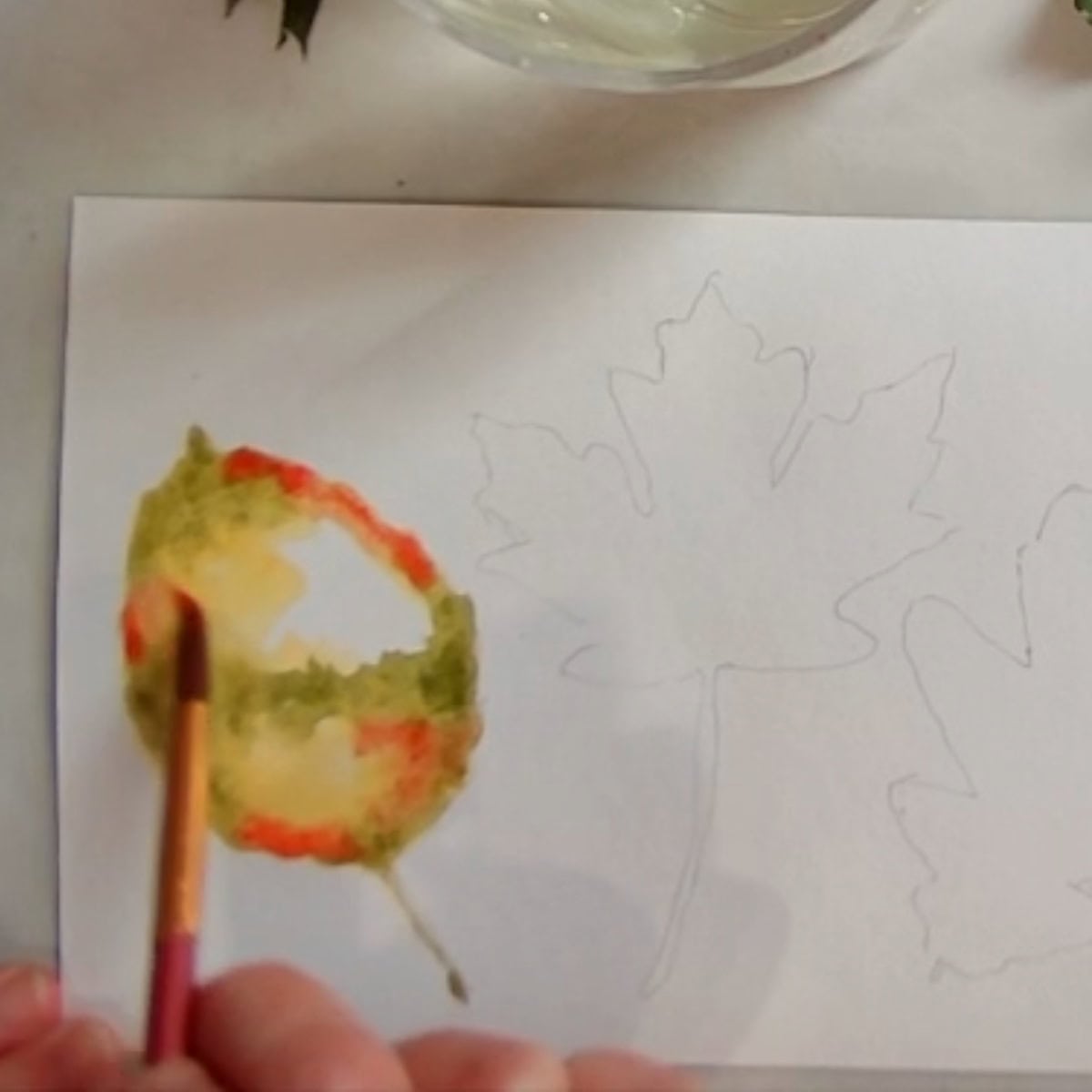 An artist painting an autumn leaf in yellow, green, and red watercolor paint.