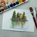 Watercolor painting of trees reflected on water next to a watercolor paint set and paintbrushes.