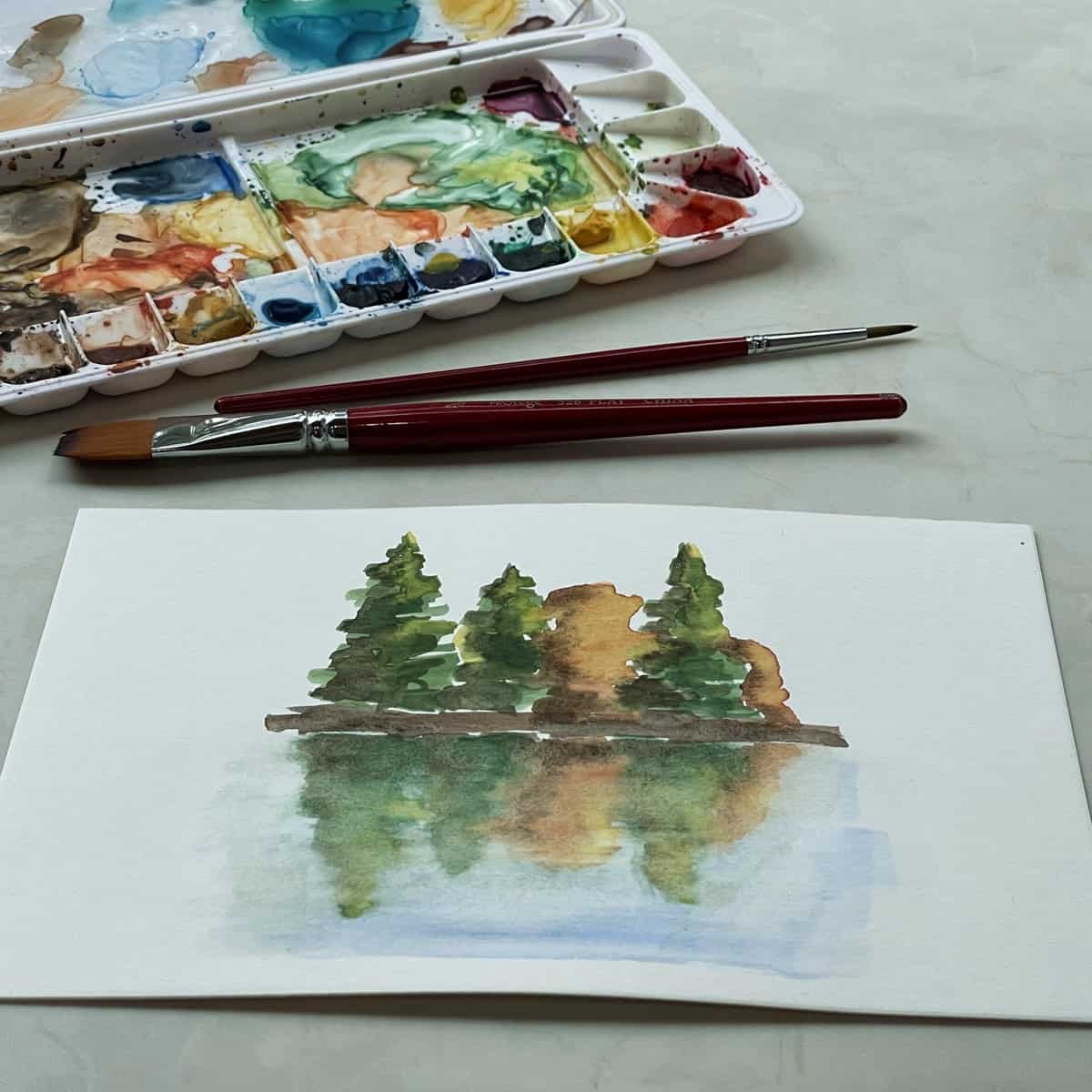 Watercolor painting of trees reflected on water next to paintbrushes and a watercolor paint set.