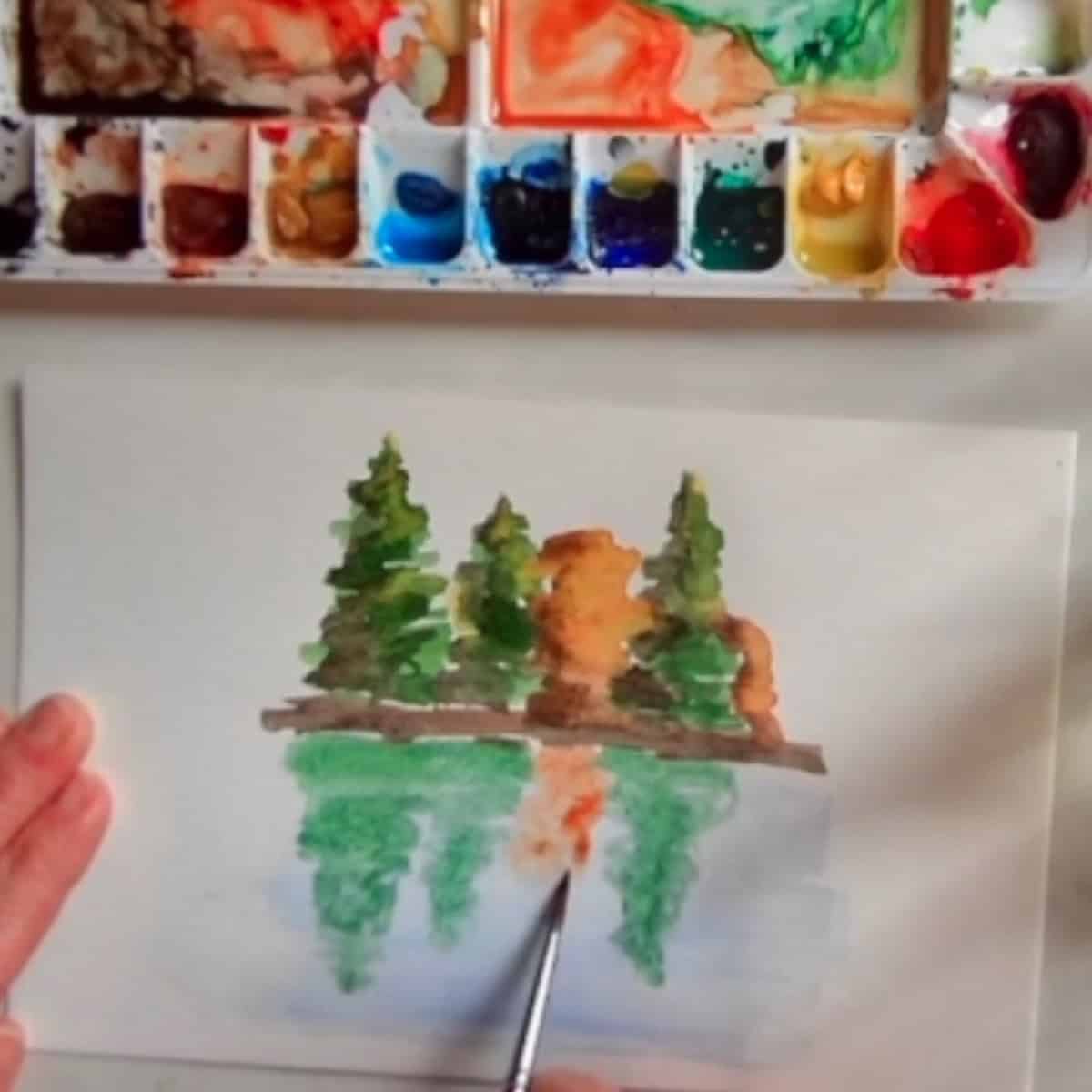The artist's hand paints in a reflection of the trees into the water below.
