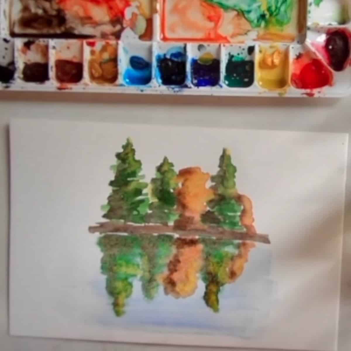 The completed painting of trees reflected on the water is displayed on a white table next to a watercolor paint set.