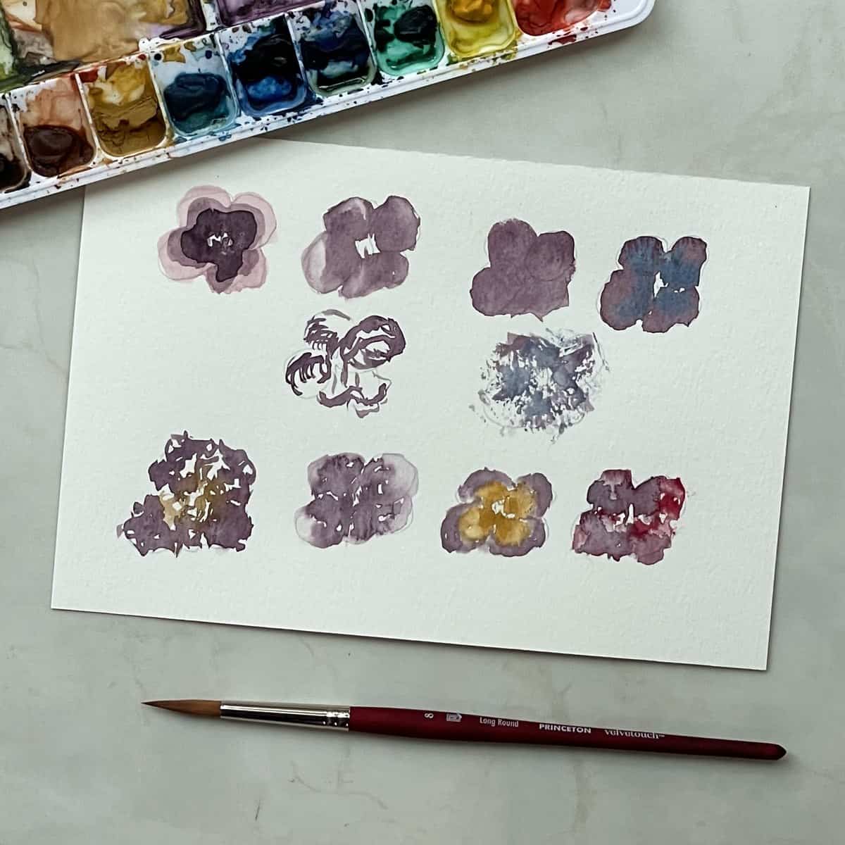 Watercolor painting of pansies that show examples of watercolor painting techniques for beginners next to a palette and paintbrush.