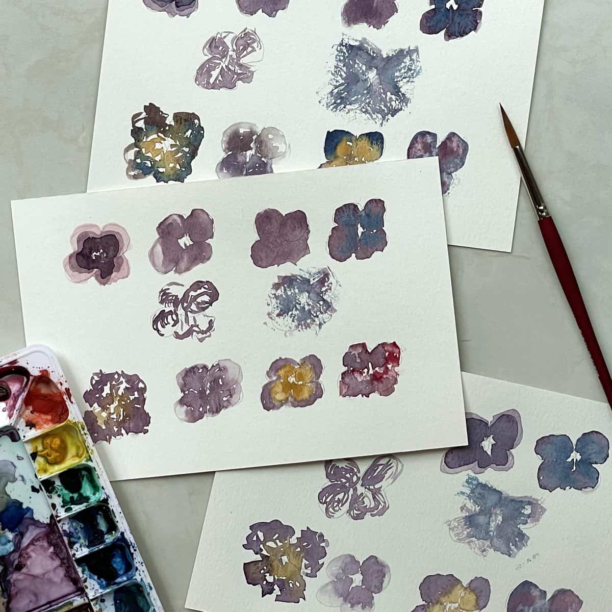 Watercolor paintings of pansies that show examples of watercolor painting techniques for beginners next to a palette and paintbrush.