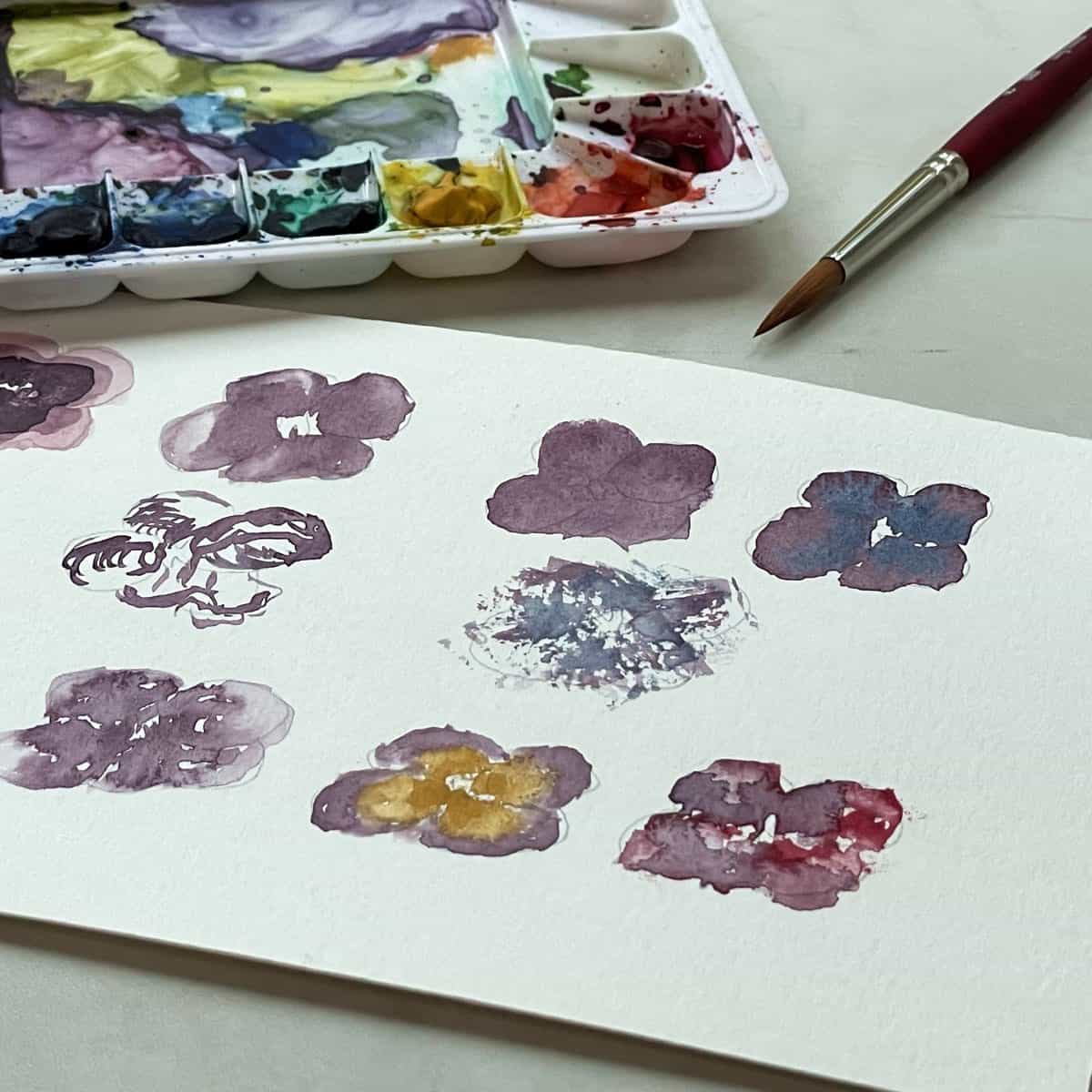 Watercolor painting of pansies that show examples of watercolor painting techniques for beginners next to a palette and paintbrush.